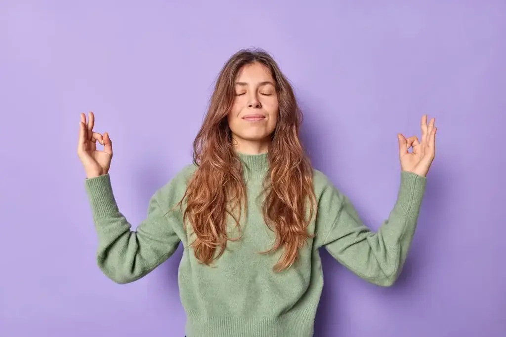 woman holds hands zen om gesture keeps eyes closed meditates indoor dressed casually keeps emotions control peace patience breathes air freely isolated purple wall 2